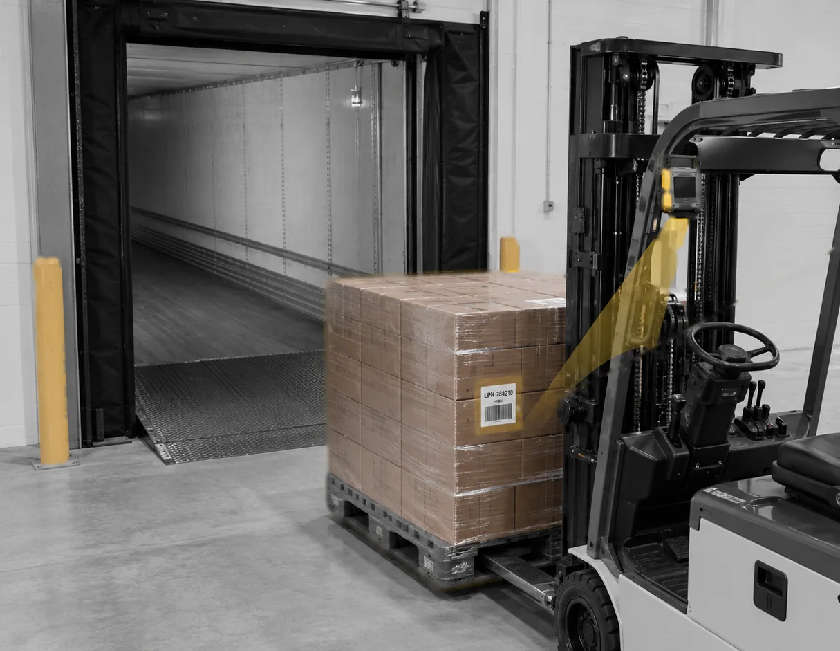 A forklift carries a pallet toward a dock door. The camera on the mast reads the pallet label.
